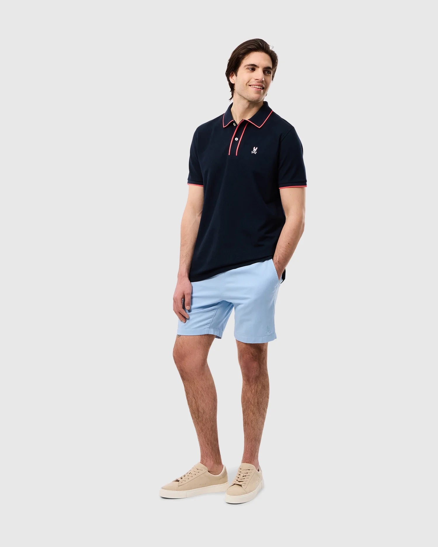 A man stands smiling, dressed in a dark polo shirt with an embroidered Psycho Bunny logo, light blue MENS WILLIS STRETCH TENCEL SHORT - B6R239Y1WB, and cream sneakers, against a plain white background.
