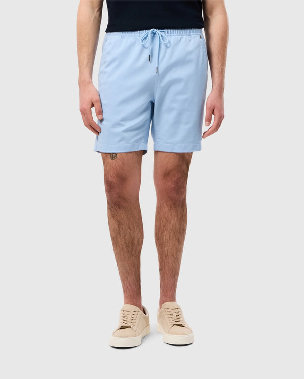 A person standing wearing light blue Psycho Bunny Stretch Tencel shorts and beige sneakers, only showing from the waist down on a white background.