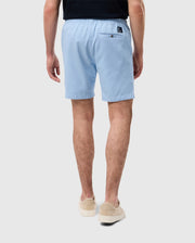 Rear view of a man wearing light blue Psycho Bunny MENS WILLIS STRETCH TENCEL SHORT - B6R239Y1WB and beige slip-on shoes, standing against a white background.