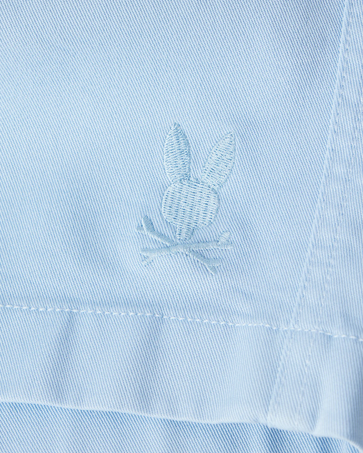 Close-up of a light blue Psycho Bunny Men's Willis Stretch Tencel Short with a textured bunny rabbit embroidered logo stitched near a seam, showing detailed embroidery in white thread.