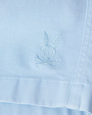 Close-up of a light blue Psycho Bunny Men's Willis Stretch Tencel Short with a textured bunny rabbit embroidered logo stitched near a seam, showing detailed embroidery in white thread.