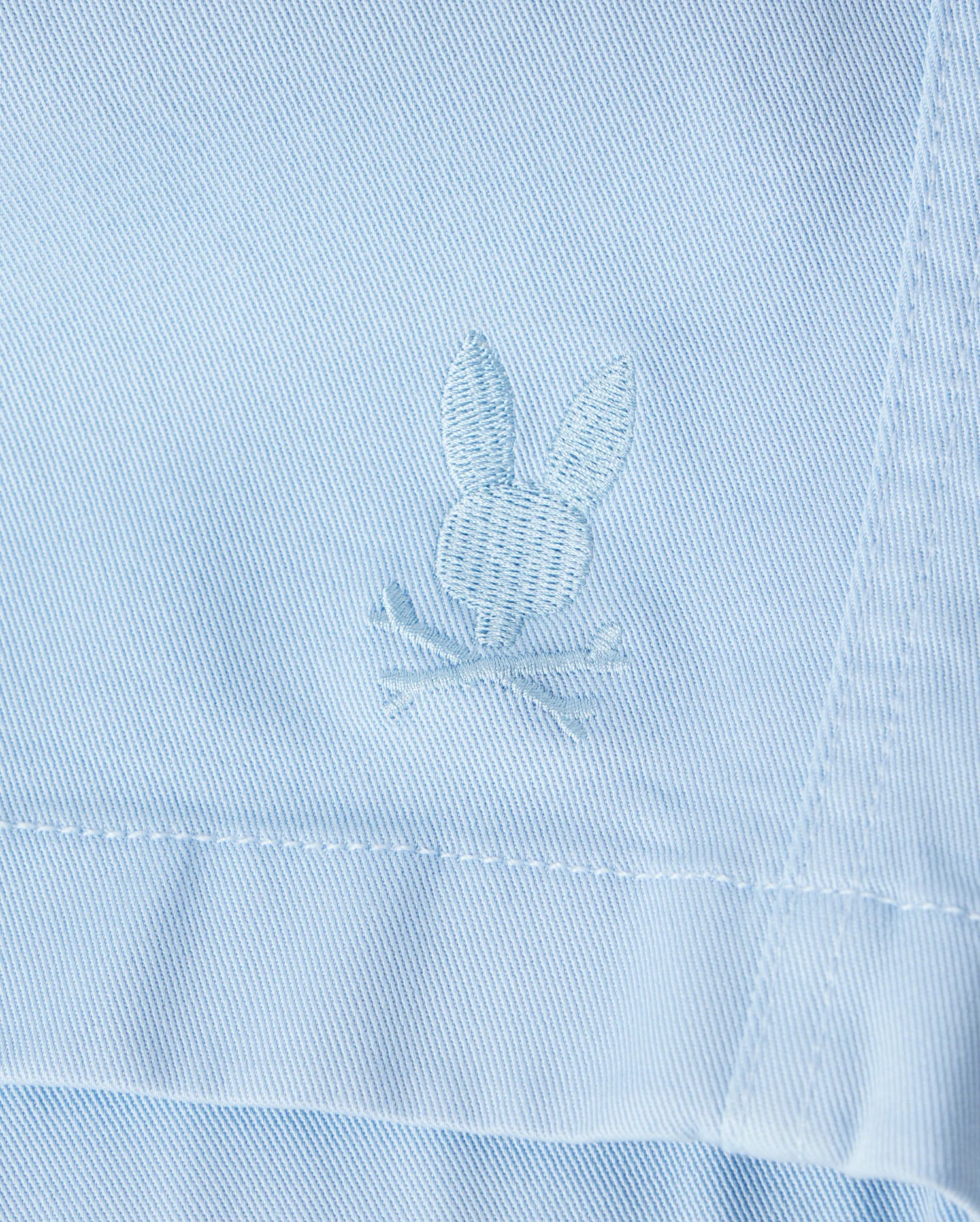 Close-up of a light blue Psycho Bunny Men's Willis Stretch Tencel Short with a textured bunny rabbit embroidered logo stitched near a seam, showing detailed embroidery in white thread.