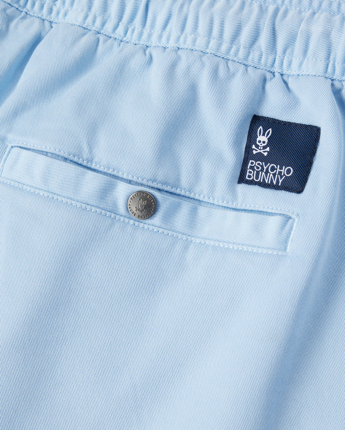 Close-up of a light blue Psycho Bunny Stretch Tencel short with a small embroidered "Psycho Bunny" logo patch and a silver button, displaying fine textile detail and neat stitching.