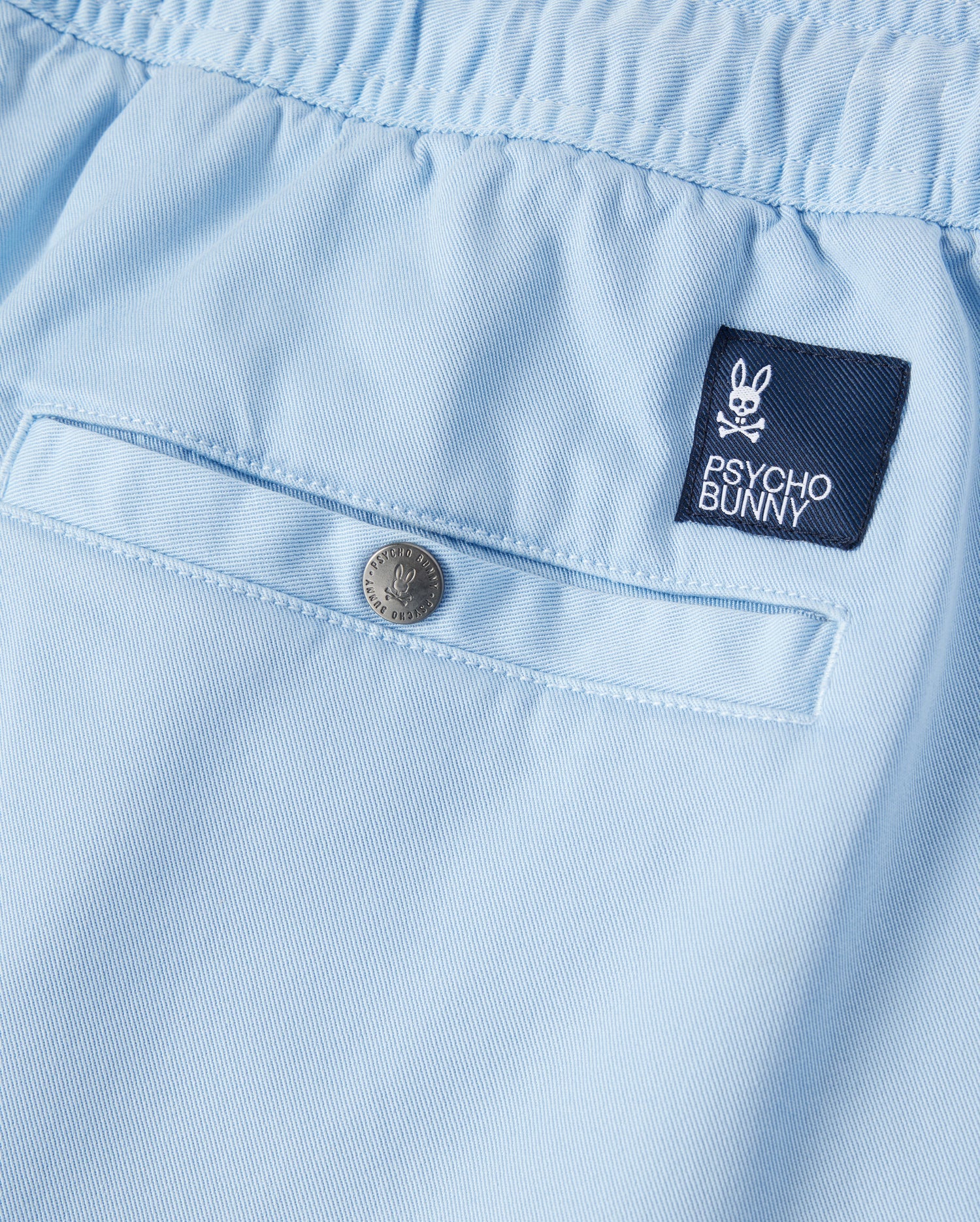 Close-up of a light blue Psycho Bunny Stretch Tencel short with a small embroidered "Psycho Bunny" logo patch and a silver button, displaying fine textile detail and neat stitching.