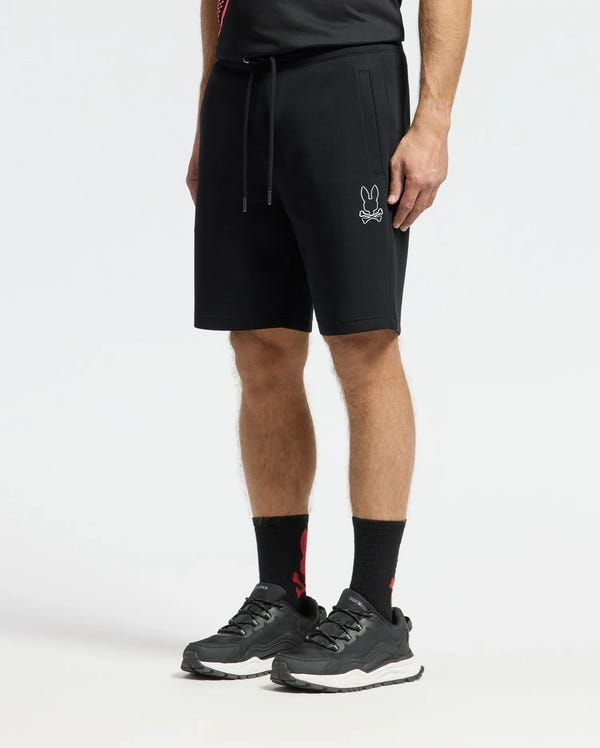 A man is wearing Psycho Bunny's MENS DACANO PERFORATED SWEATSHORT - B6R286E200 with a rabbit logo, styled for an athletic fit. He's also in black sneakers, high black socks, and a performance-oriented cotton-blend jersey shirt, all set against a plain white background.