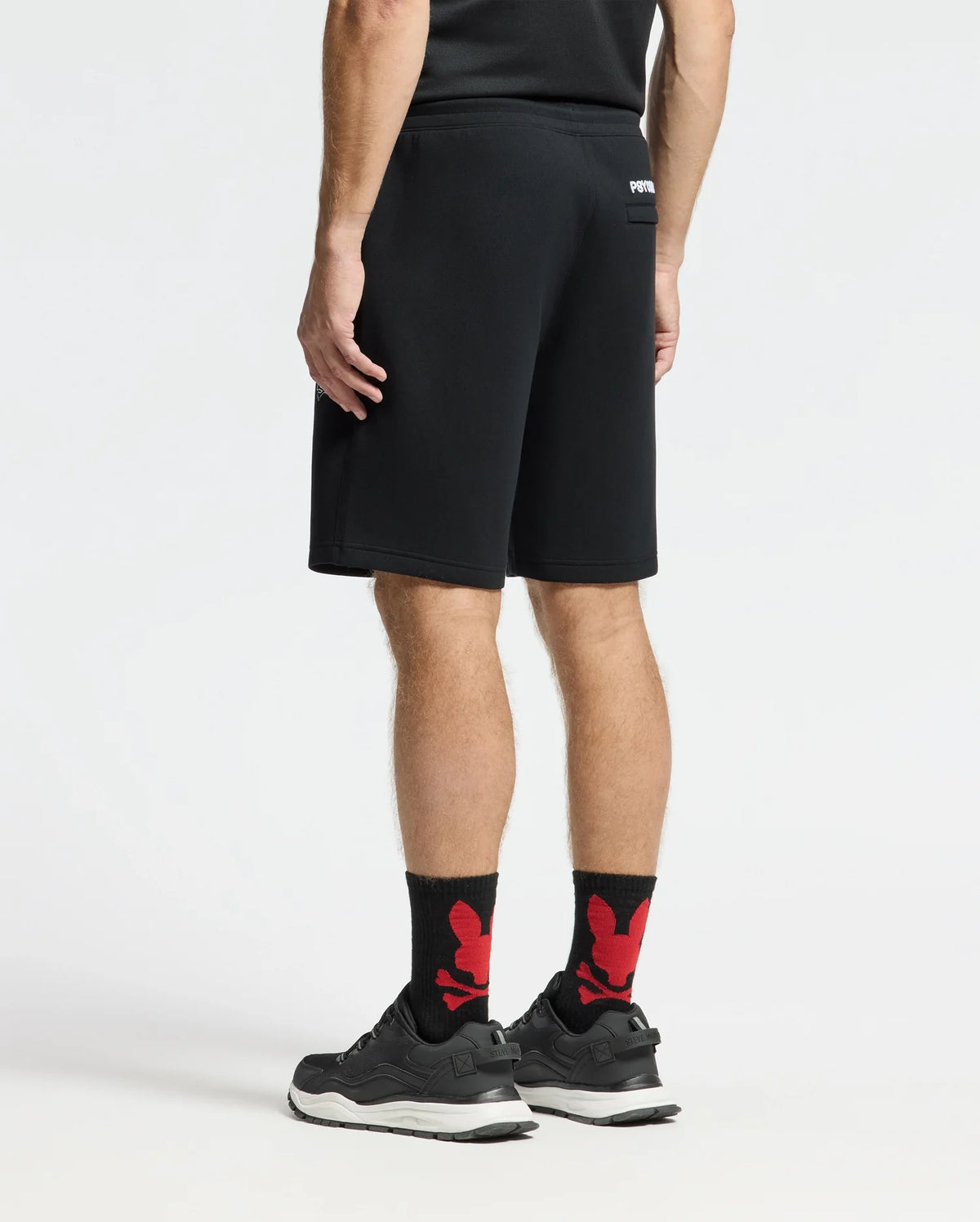 A person stands facing away, wearing a black athletic shirt and MENS DACANO PERFORATED SWEATSHORT by Psycho Bunny along with black sneakers and black socks featuring red designs. This cotton-blend jersey ensemble is performance-oriented against a plain white background.