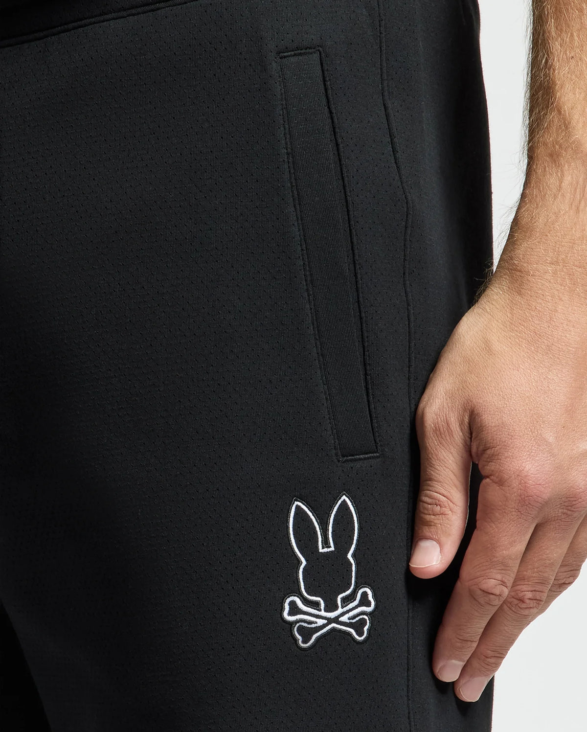 Close-up of someone wearing black MENS DACANO PERFORATED SWEATSHORT by Psycho Bunny. The cotton-blend jersey features a white bunny head with crossed bones logo near the knee. Their hand rests by their side, exuding casual confidence.