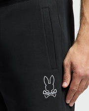 Close-up of someone wearing black MENS DACANO PERFORATED SWEATSHORT by Psycho Bunny. The cotton-blend jersey features a white bunny head with crossed bones logo near the knee. Their hand rests by their side, exuding casual confidence.