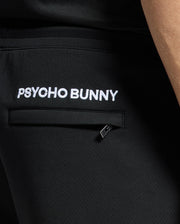 A person wears MENS DACANO PERFORATED SWEATSHORT by Psycho Bunny, featuring "PSYCHO BUNNY" embroidered in white above the back pocket and a logo tag nearby, highlighting its performance design.