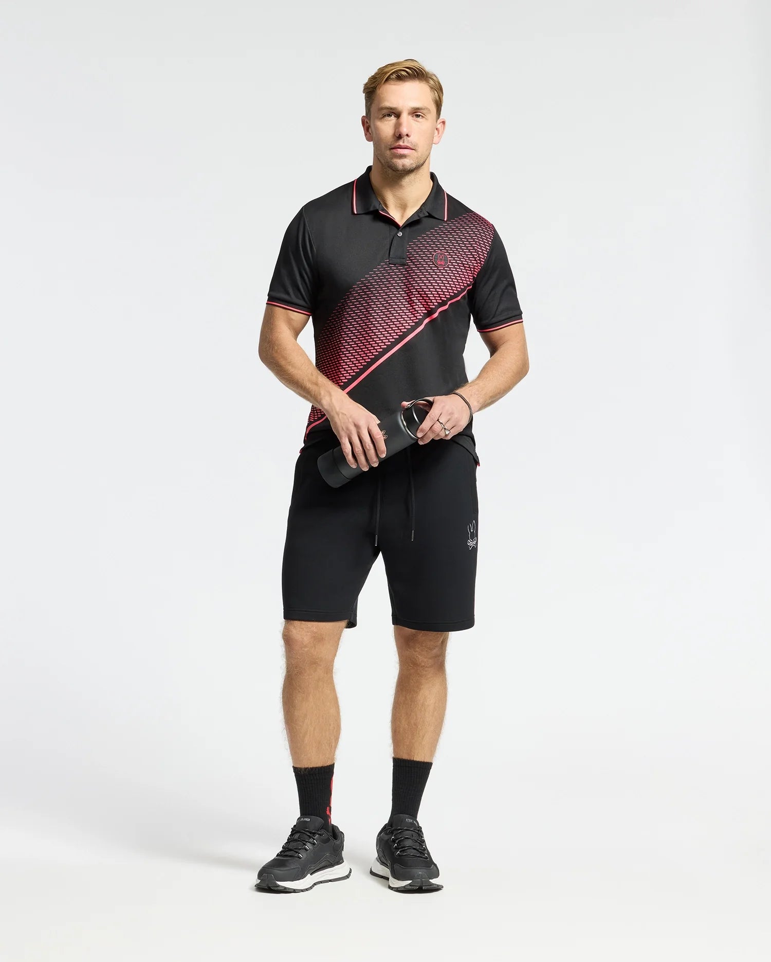 A man in Psycho Bunny's black and red patterned polo shirt made of a cotton-blend jersey and MENS DACANO PERFORATED SWEATSHORT stands holding a water bottle. He has an athletic fit, wearing sleek black running shoes and socks against a white background.