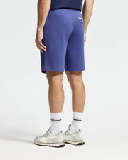 A person faces away from the camera, wearing Psycho Bunny's Dacono Perforated Sweatshorts and a navy performance cotton-blend shirt, paired with white socks and gray sneakers, creating an athletic look against a plain white background.