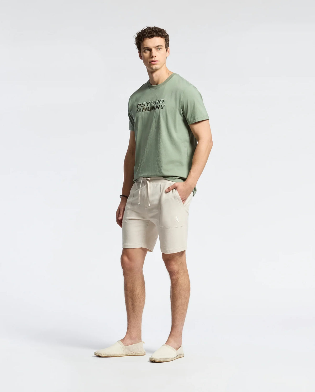 Wearing Psycho Bunny's MENS TAYLOR LIGHT PIQUE SWEATSHORT - B6R344F200 in beige, a person sports a lightweight light green Pima cotton t-shirt with text and slip-on shoes. They stand against a plain white backdrop, showcasing curly hair and a casual-chic look as they pose relaxedly at the camera.