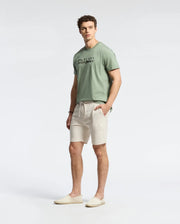 Wearing Psycho Bunny's MENS TAYLOR LIGHT PIQUE SWEATSHORT - B6R344F200 in beige, a person sports a lightweight light green Pima cotton t-shirt with text and slip-on shoes. They stand against a plain white backdrop, showcasing curly hair and a casual-chic look as they pose relaxedly at the camera.