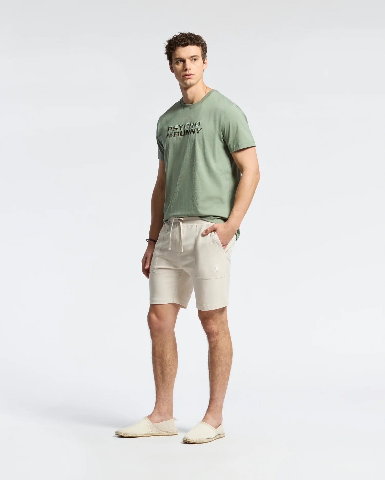 Wearing Psycho Bunny's MENS TAYLOR LIGHT PIQUE SWEATSHORT - B6R344F200 in beige, a person sports a lightweight light green Pima cotton t-shirt with text and slip-on shoes. They stand against a plain white backdrop, showcasing curly hair and a casual-chic look as they pose relaxedly at the camera.