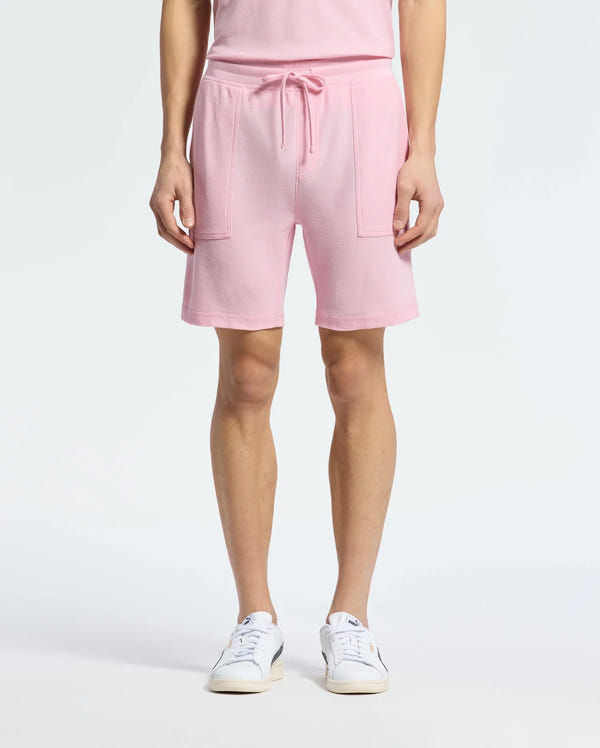 A person wearing Psycho Bunny's MENS TAYLOR LIGHT PIQUE SWEATSHORT in pink, featuring a drawstring waistband and patch pockets, paired with a matching top, embodies casual chic. The look is completed with white sneakers on a plain white background.