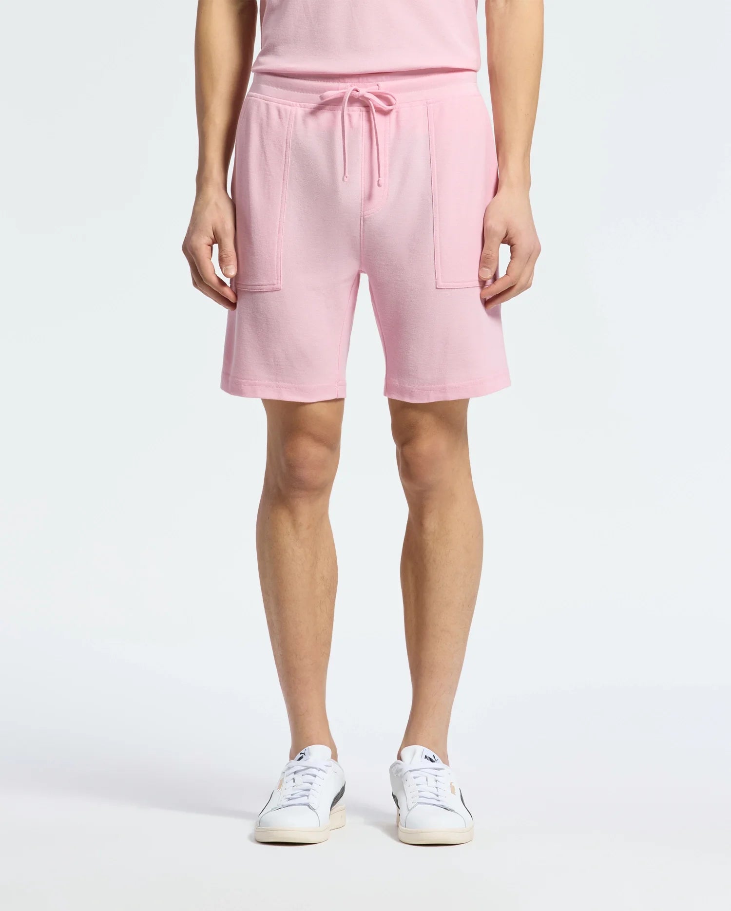 A person wearing Psycho Bunny's MENS TAYLOR LIGHT PIQUE SWEATSHORT in pink, featuring a drawstring waistband and patch pockets, paired with a matching top, embodies casual chic. The look is completed with white sneakers on a plain white background.