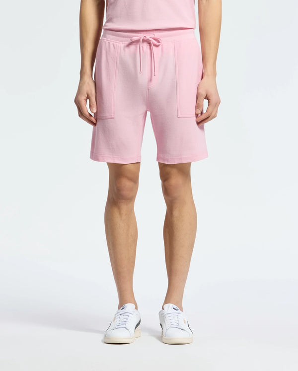 A person wearing Psycho Bunny's MENS TAYLOR LIGHT PIQUE SWEATSHORT in pink, featuring a drawstring waistband and patch pockets, paired with a matching top, embodies casual chic. The look is completed with white sneakers on a plain white background.
