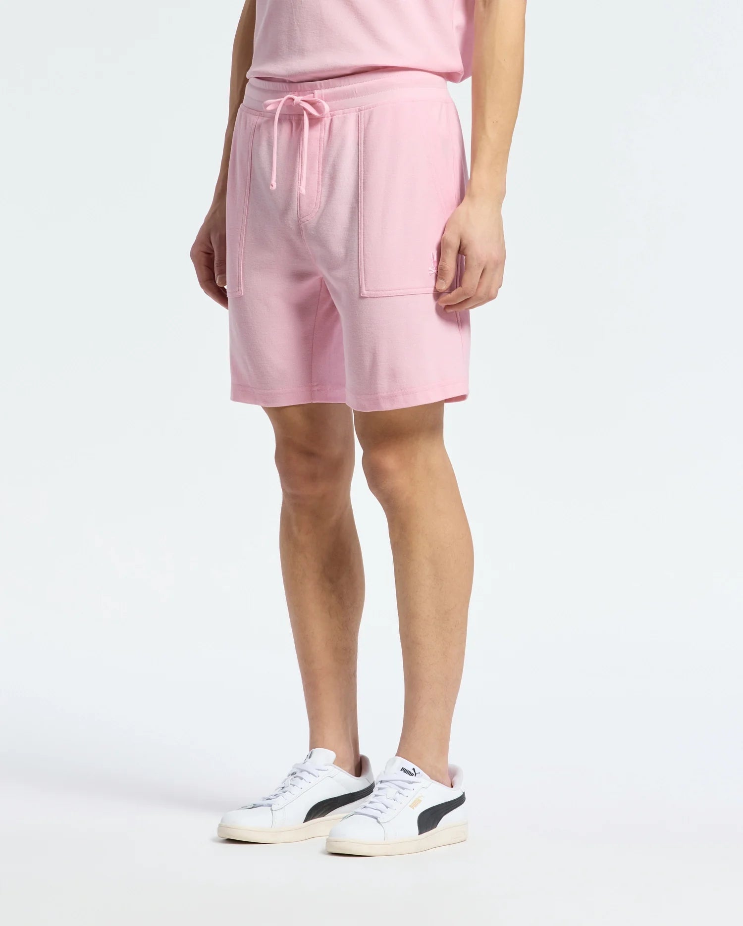A person in a casual-chic wardrobe wears the MENS TAYLOR LIGHT PIQUE SWEATSHORT - B6R344F200 by Psycho Bunny, crafted from lightweight piqué material, paired with a matching pink T-shirt. The shorts feature a drawstring and large pockets, and are styled with white sneakers highlighted by black details.