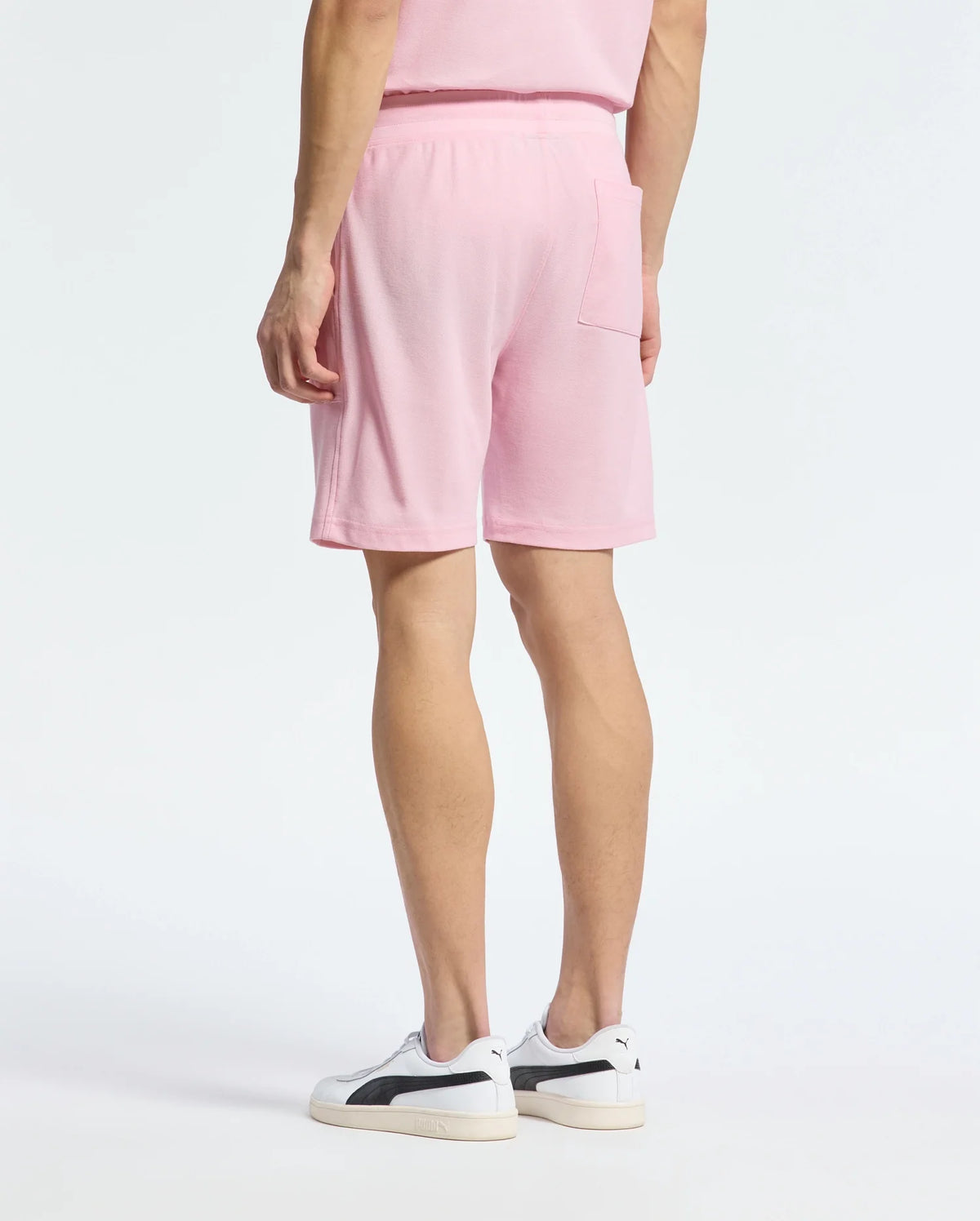 A person stands with their back to the viewer in a casual-chic Psycho Bunny outfit, wearing a pink T-shirt and MENS TAYLOR LIGHT PIQUE SWEATSHORT - B6R344F200. White sneakers with black accents complete the look against a plain white background, showcasing the lightweight piqué fabric's comfort.