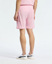 A person stands with their back to the viewer in a casual-chic Psycho Bunny outfit, wearing a pink T-shirt and MENS TAYLOR LIGHT PIQUE SWEATSHORT - B6R344F200. White sneakers with black accents complete the look against a plain white background, showcasing the lightweight piqué fabric's comfort.