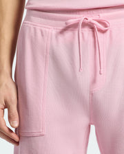 A close-up shows a person in Psycho Bunny's pink drawstring sweatshorts and matching top, showcasing casual-chic style. The soft, textured fabric with visible seams highlights the left hand by their side.