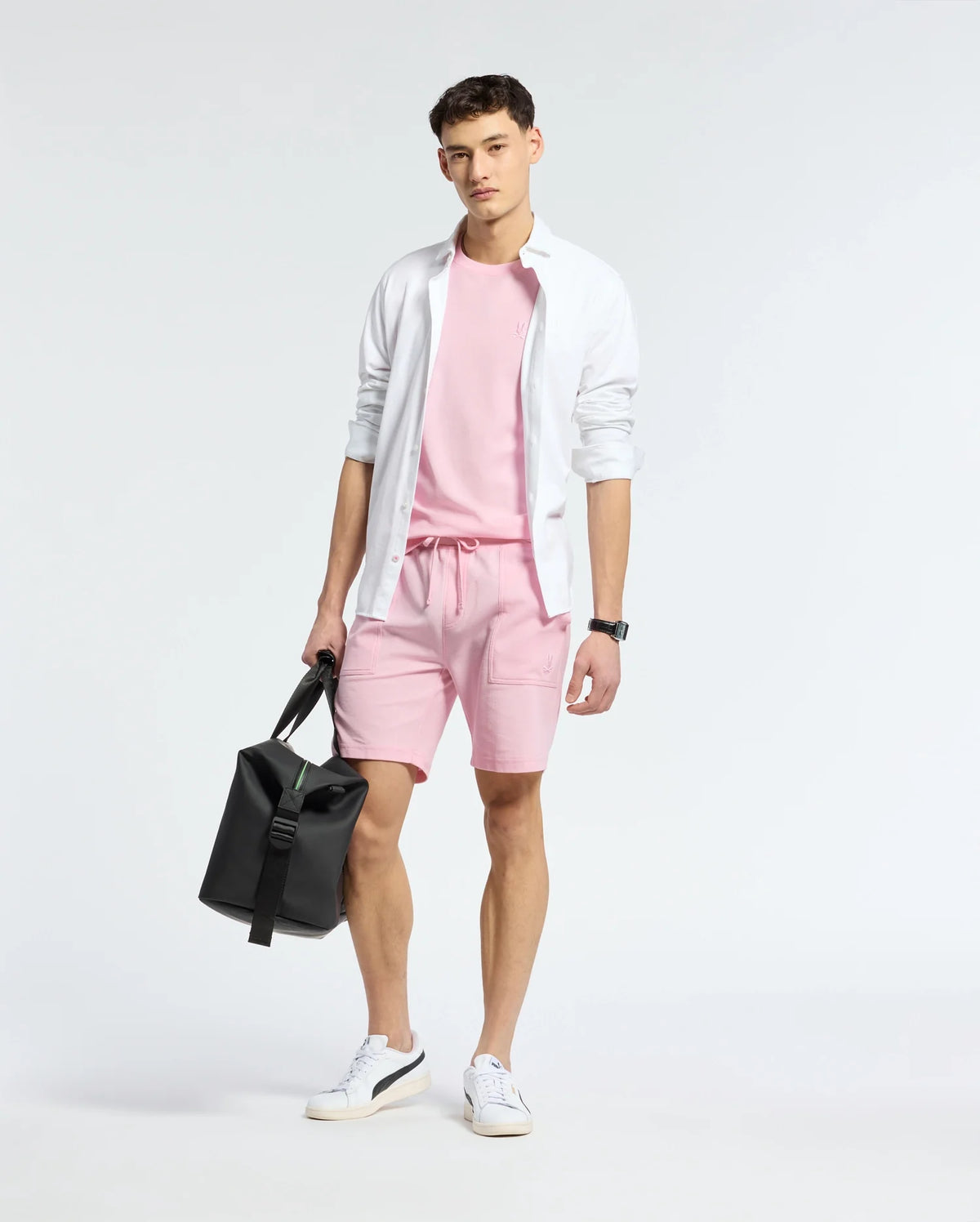 In front of a simple backdrop, an individual showcases a casual-chic outfit featuring a white shirt, a pink T-shirt, and Psycho Bunny's MENS TAYLOR LIGHT PIQUE SWEATSHORT. They accessorize with a black bag and white sneakers.