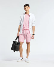 In front of a simple backdrop, an individual showcases a casual-chic outfit featuring a white shirt, a pink T-shirt, and Psycho Bunny's MENS TAYLOR LIGHT PIQUE SWEATSHORT. They accessorize with a black bag and white sneakers.