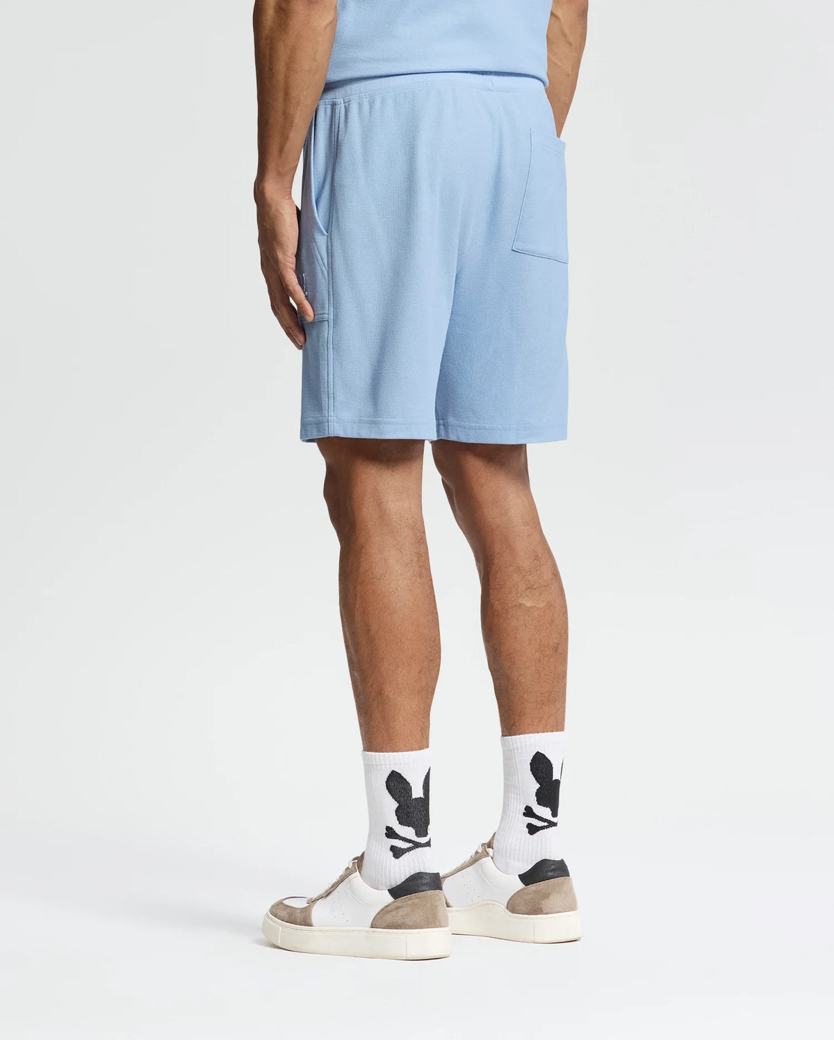 Wearing Psycho Bunny's Taylor Light Pique Sweatshorts in light blue, a coordinated shirt, white socks with black designs, and beige-white sneakers, the individual presents an effortlessly stylish look against a plain white background—ideal for a casual-chic wardrobe.