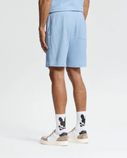 Wearing Psycho Bunny's Taylor Light Pique Sweatshorts in light blue, a coordinated shirt, white socks with black designs, and beige-white sneakers, the individual presents an effortlessly stylish look against a plain white background—ideal for a casual-chic wardrobe.