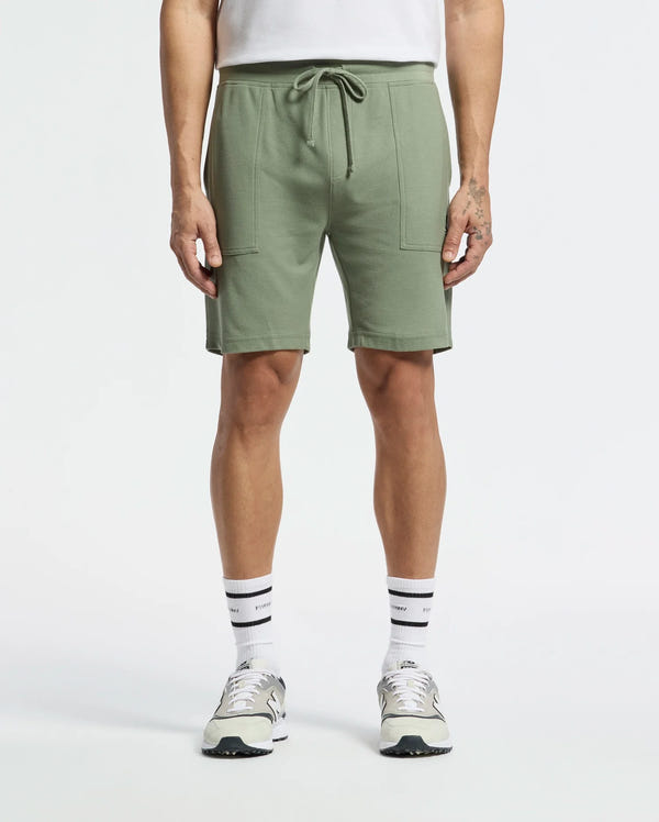 Sporting a casual-chic look, the person wears Psycho Bunny's MENS TAYLOR LIGHT PIQUE SWEATSHORT - B6R344F200 in light green with a drawstring and large front pockets, paired with white socks, gray and white sneakers, and a tucked-in white shirt against a plain white background.