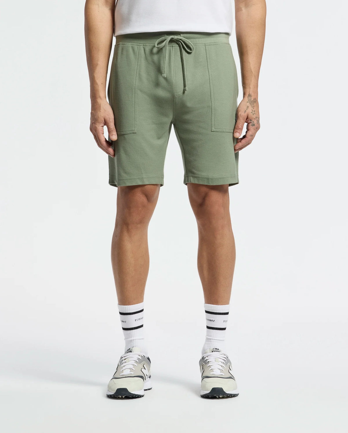Sporting a casual-chic look, the person wears Psycho Bunny's MENS TAYLOR LIGHT PIQUE SWEATSHORT - B6R344F200 in light green with a drawstring and large front pockets, paired with white socks, gray and white sneakers, and a tucked-in white shirt against a plain white background.