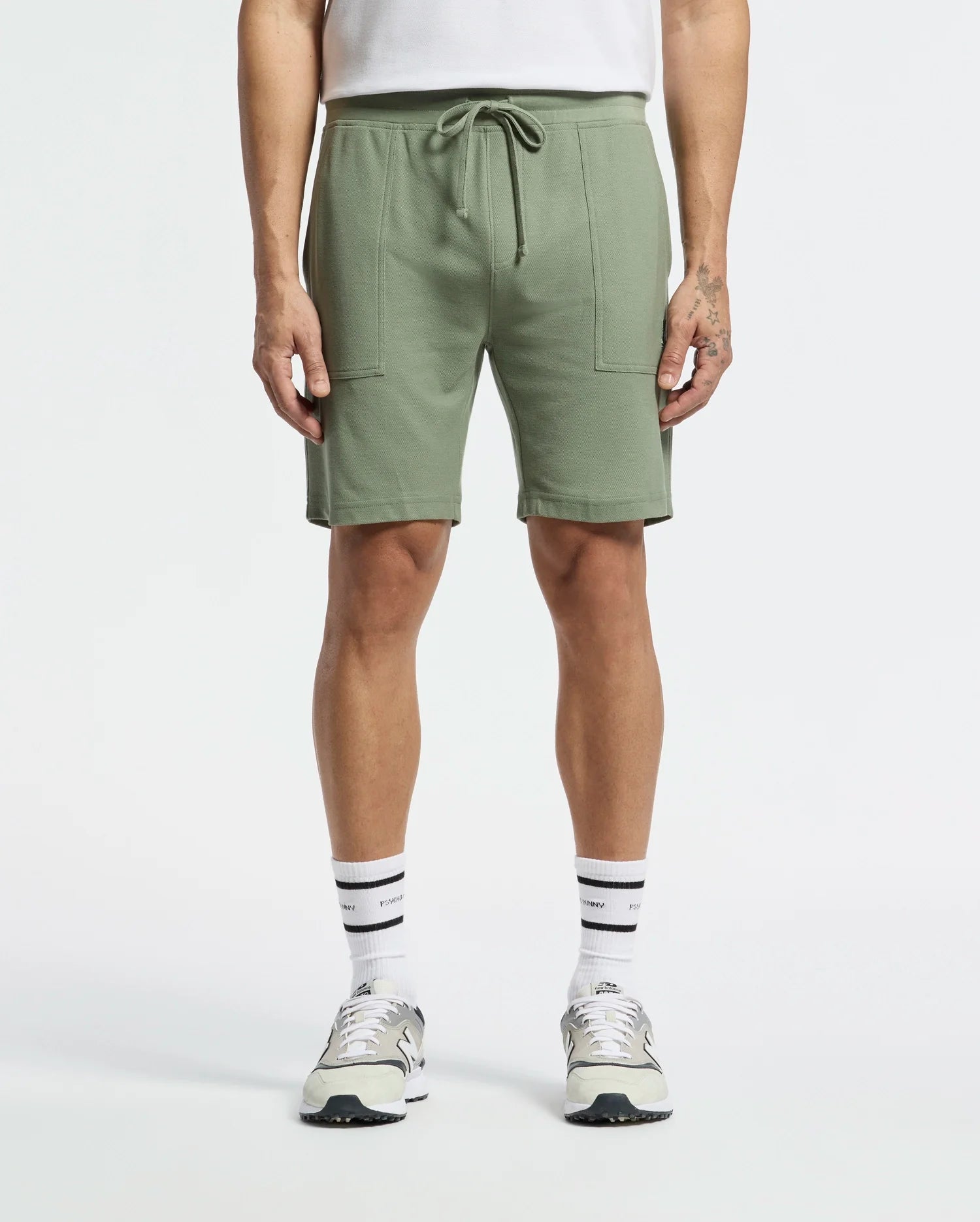 Sporting a casual-chic look, the person wears Psycho Bunny's MENS TAYLOR LIGHT PIQUE SWEATSHORT - B6R344F200 in light green with a drawstring and large front pockets, paired with white socks, gray and white sneakers, and a tucked-in white shirt against a plain white background.