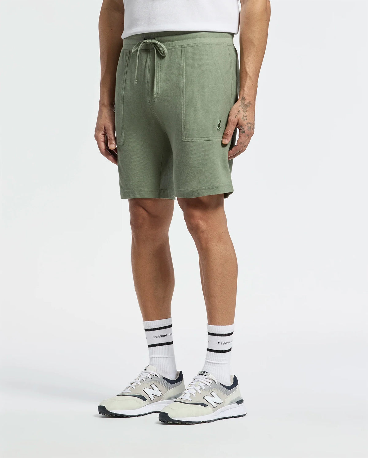 A person exudes casual-chic style wearing Psycho Bunny's MENS TAYLOR LIGHT PIQUE SWEATSHORT, paired with gray and white sneakers, black-striped socks, and a white Pima cotton piqué shirt. Their hands rest by their sides, highlighting a wrist tattoo against the plain white backdrop.