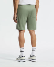 A person is seen from the back in Psycho Bunny's MENS TAYLOR LIGHT PIQUE SWEATSHORT - B6R344F200 in green, paired with a white Pima cotton piqué shirt, white socks, and sneakers. Their left hand hangs by their side, and a back pocket appears on the shorts' right against a plain white backdrop for a casual-chic look.