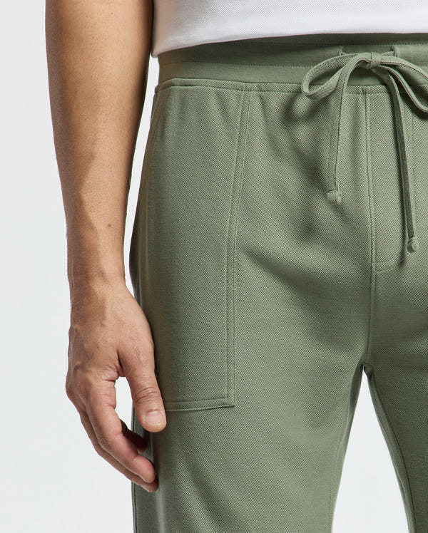 Close-up of a person wearing Psycho Bunny's MENS TAYLOR LIGHT PIQUE SWEATSHORT in light green with a white top, highlighting the hand near the pocket and the texture of Pima cotton piqué fabric.