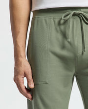 Close-up of a person wearing Psycho Bunny's MENS TAYLOR LIGHT PIQUE SWEATSHORT in light green with a white top, highlighting the hand near the pocket and the texture of Pima cotton piqué fabric.
