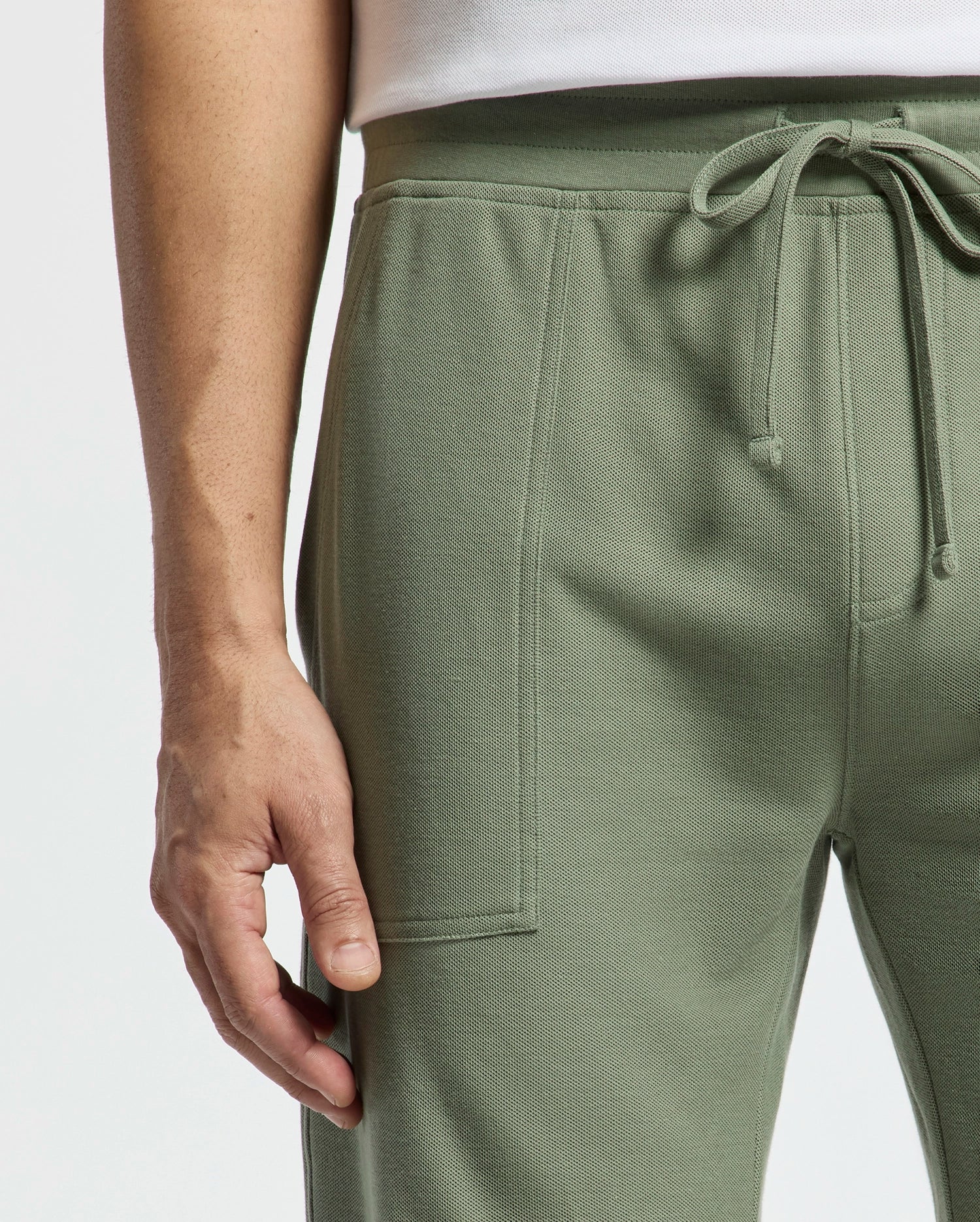 Close-up of a person wearing Psycho Bunny's MENS TAYLOR LIGHT PIQUE SWEATSHORT in light green with a white top, highlighting the hand near the pocket and the texture of Pima cotton piqué fabric.