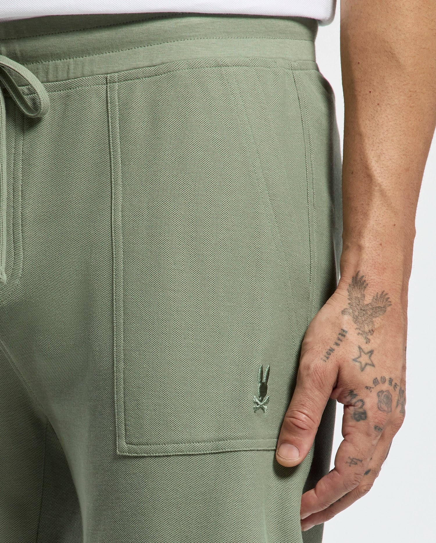 A person wears Psycho Bunny MENS TAYLOR LIGHT PIQUE SWEATSHORT in sage green Pima cotton, featuring a small embroidered logo by the pocket. Their tattooed hand rests nearby, while an elegant white top complements the casual-chic look.
