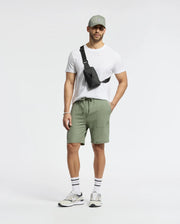 A man in a light olive Psycho Bunny cap, sneakers, and MENS TAYLOR LIGHT PIQUE SWEATSHORT by Psycho Bunny pairs them with a white t-shirt, black crossbody bag, sports bracelet, and black-and-white striped socks—ideal for the casual-chic enthusiast.