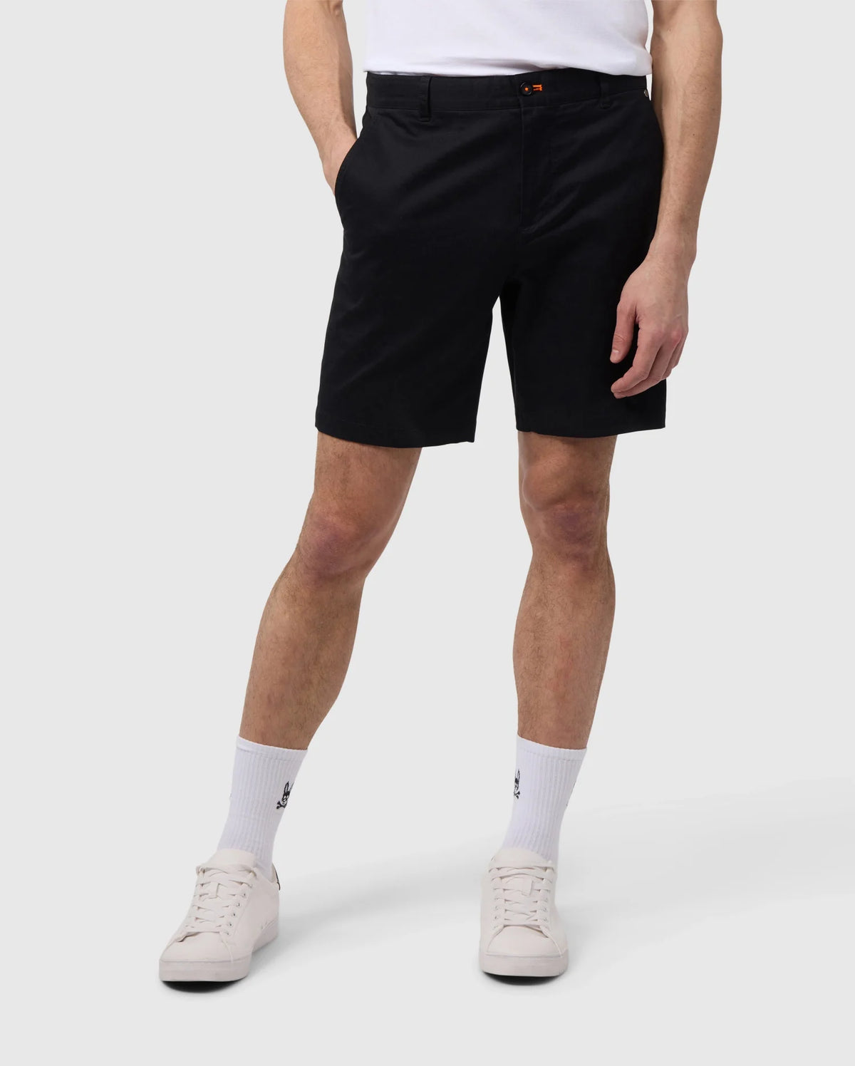 Man wearing Psycho Bunny black elastic waist shorts, white socks with logo, and white sneakers, standing against a plain background, focusing on the lower half of the body.