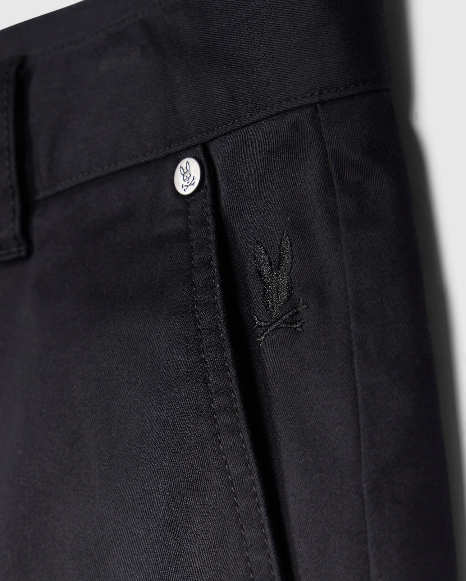 Close-up view of a black trouser waistline featuring a button with the Psycho Bunny logo and a small embroidered Psycho Bunny logo on the front pocket of men's MENS YORK CHINO SHORT - B6R357B200.