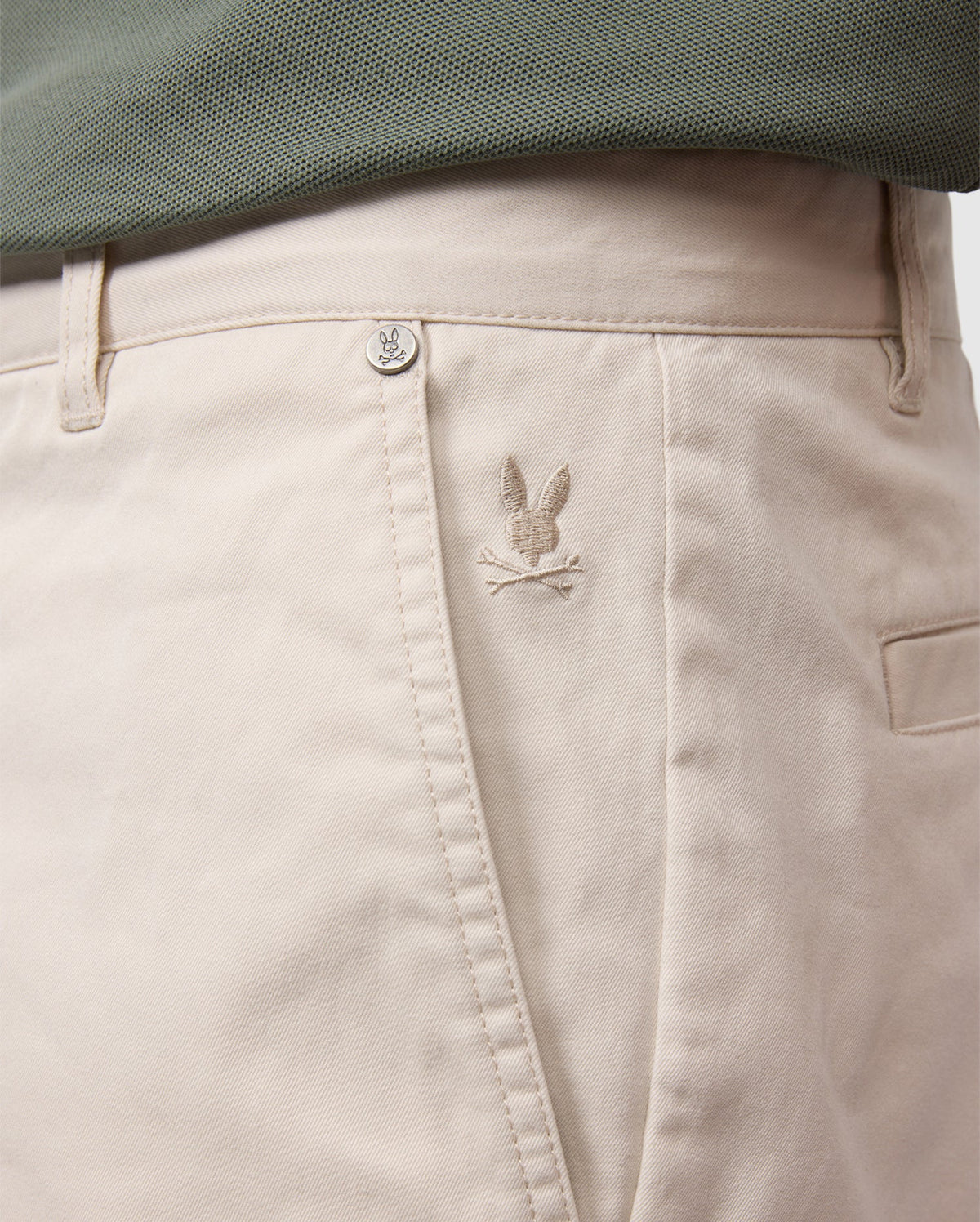 Close-up of light beige Psycho Bunny men's York chino shorts with a small, embroidered bird logo near the right front pocket, partially covered by a teal fabric edge.