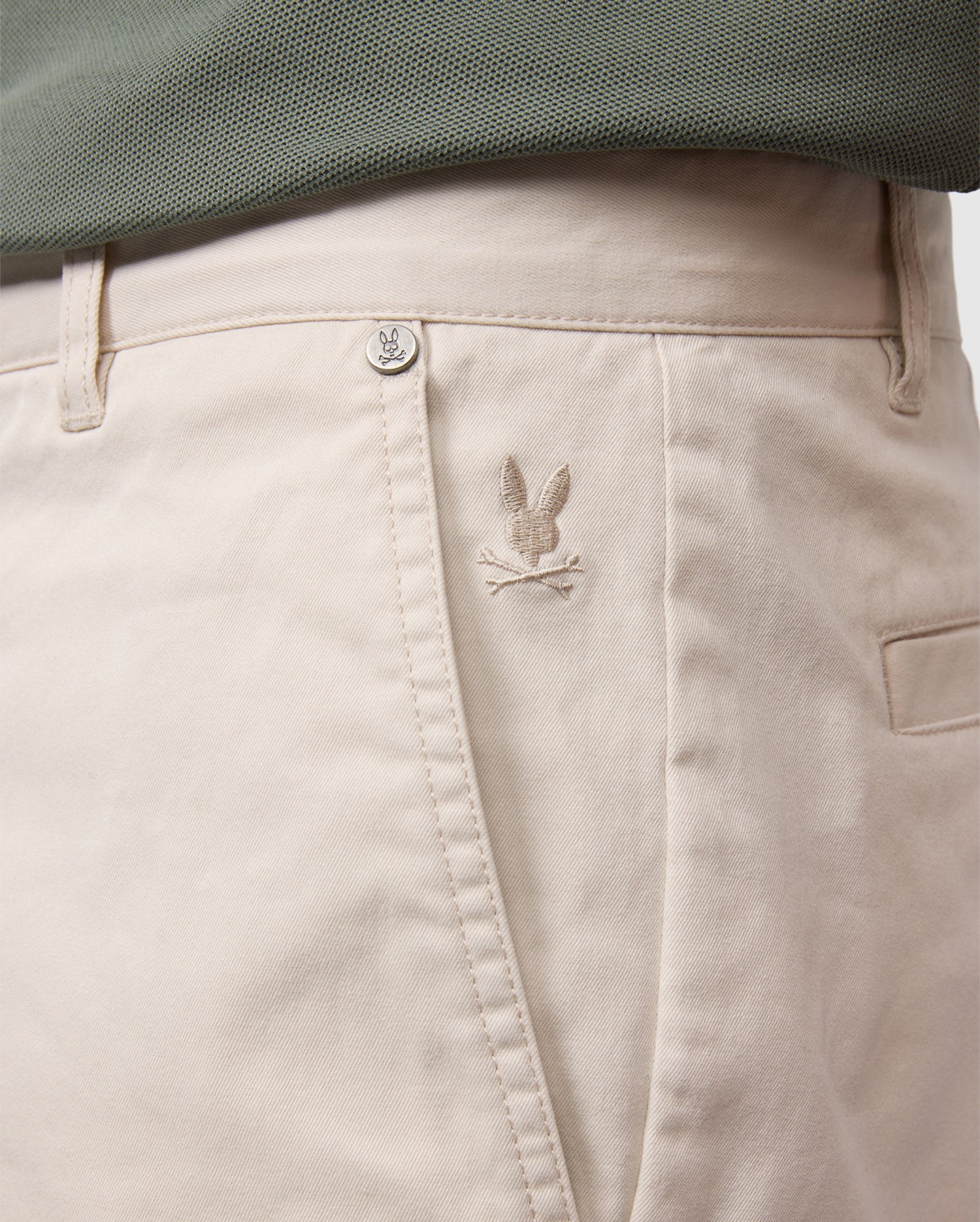 Close-up of light beige Psycho Bunny men's York chino shorts with a small, embroidered bird logo near the right front pocket, partially covered by a teal fabric edge.