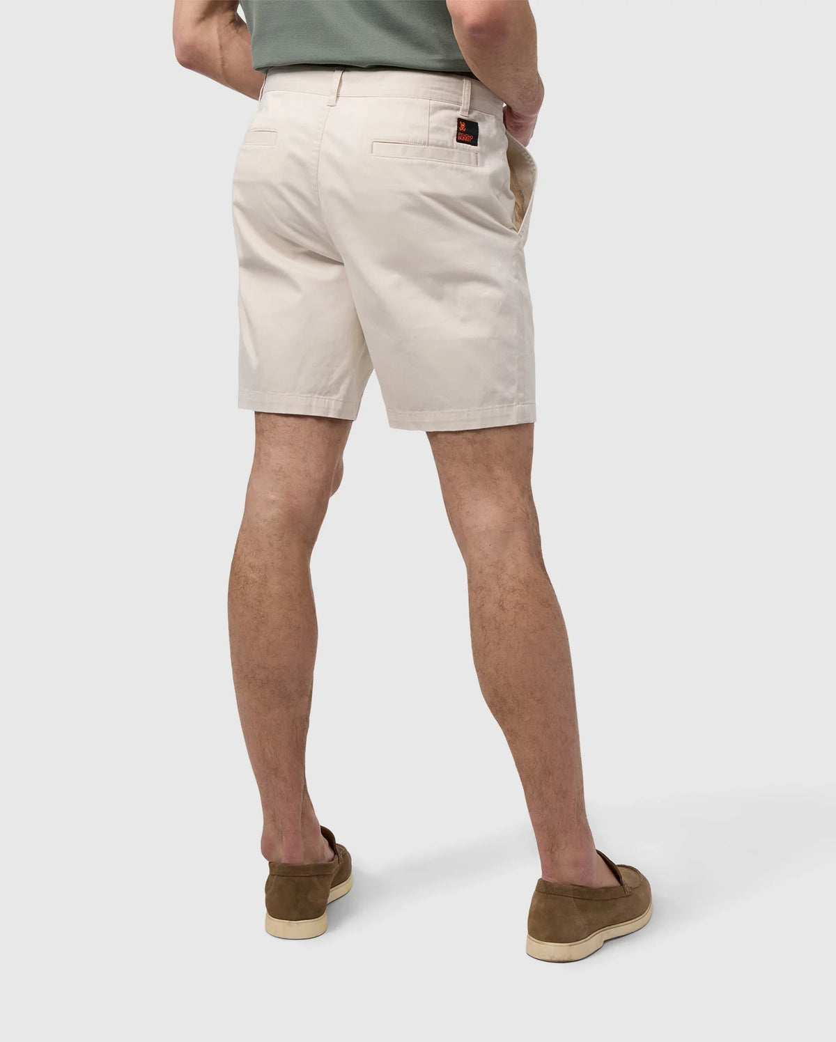 A man viewed from behind wearing Psycho Bunny's MENS YORK CHINO SHORT - B6R357B200 and brown loafers against a plain white background. His left hand is in his pocket, and he appears to be standing casually.