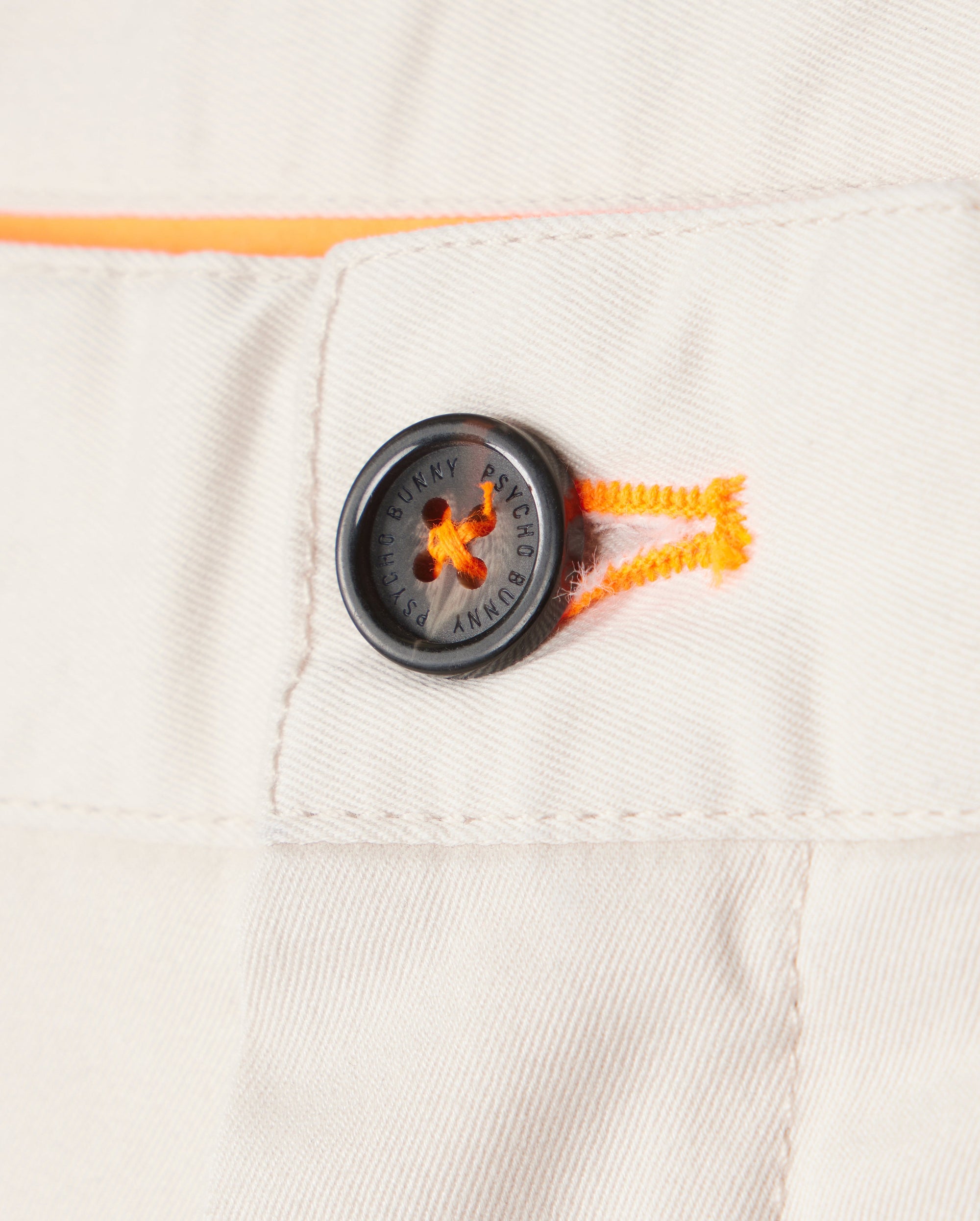Close-up of a black button on white Psycho Bunny Men's York Chino Shorts with distinctive orange stitching detail and embossed text. The button is securely sewn to the garment.