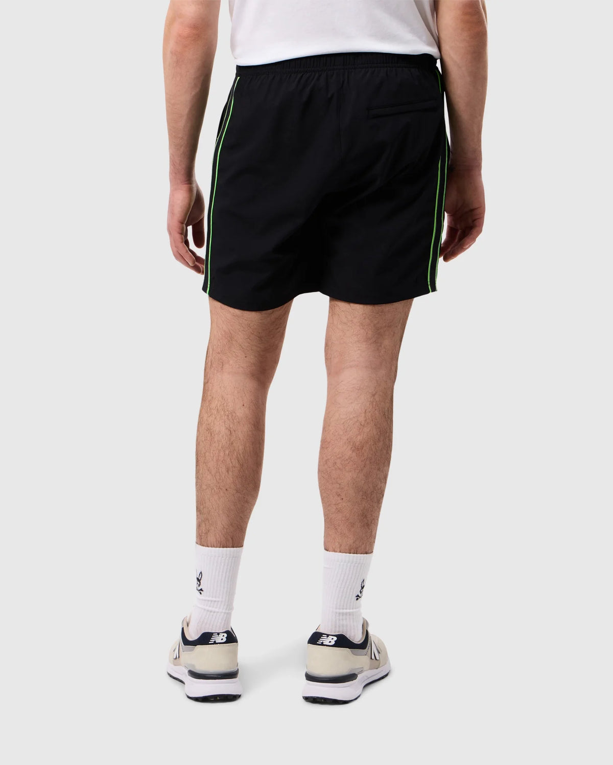 A person stands facing away from the camera, dressed in a white t-shirt and quick-drying, black Psycho Bunny MENS SONTERRA STRIPE SPORT SHORT - B6R464C200 with green side stripes. White socks and New Balance sneakers complete the look, set against a plain, light-colored background.