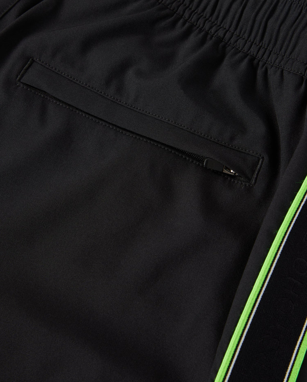 Close-up image of black athletic pants made from breathable fabric, showing a zippered back pocket detail and a green stripe running along the side. The waistband appears elastic, providing a comfortable fit. This is the MENS SONTERRA STRIPE SPORT SHORT - B6R464C200 by Psycho Bunny.