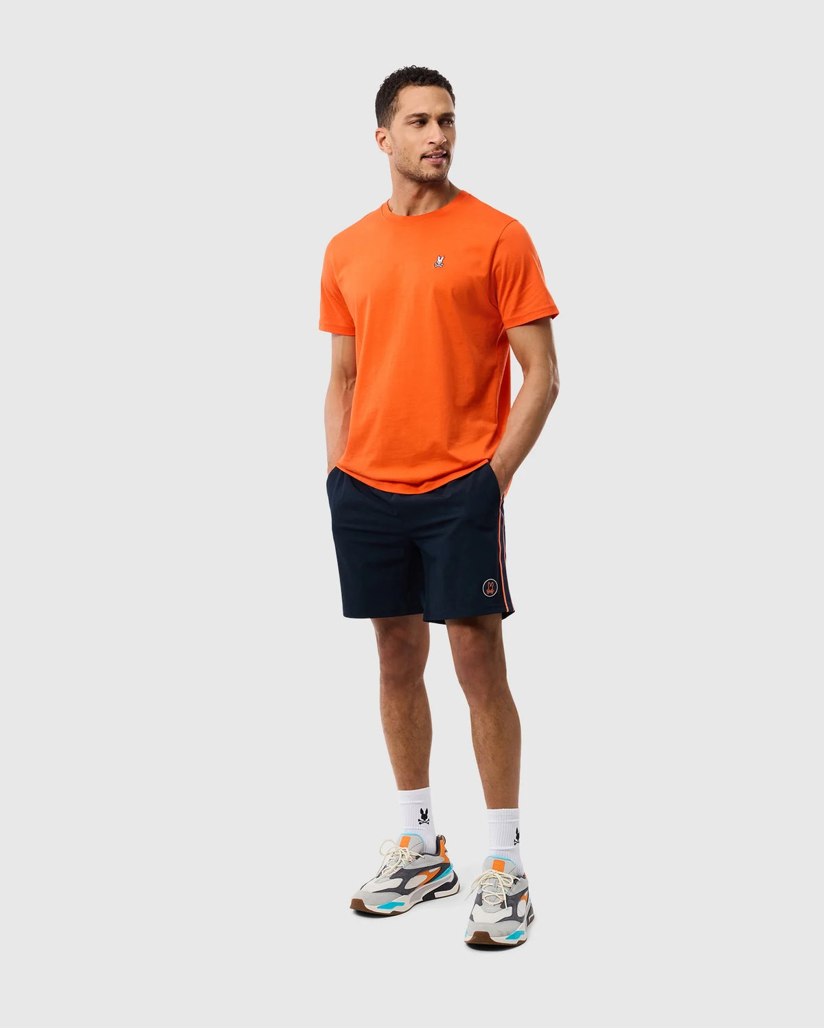 A man stands with his hands in his pockets, wearing an orange T-shirt, Psycho Bunny MENS SONTERRA STRIPE SPORT SHORT - B6R464C200 made of stretch fabric, white socks, and multicolored sneakers. He has short hair and a short beard. The background is plain and light gray.