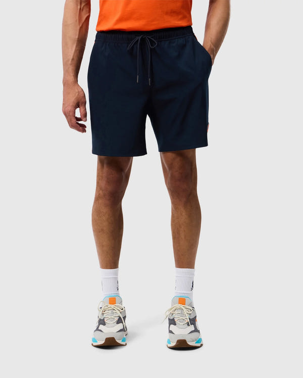 A person is standing against a plain background, wearing a bright orange t-shirt, dark blue Psycho Bunny MENS SONTERRA STRIPE SPORT SHORT - B6R464C200 with a drawstring made from stretch fabric, white socks, and multicolored athletic shoes. The individual is visible from the shoulders down.