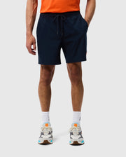 A person is standing against a plain background, wearing a bright orange t-shirt, dark blue Psycho Bunny MENS SONTERRA STRIPE SPORT SHORT - B6R464C200 with a drawstring made from stretch fabric, white socks, and multicolored athletic shoes. The individual is visible from the shoulders down.